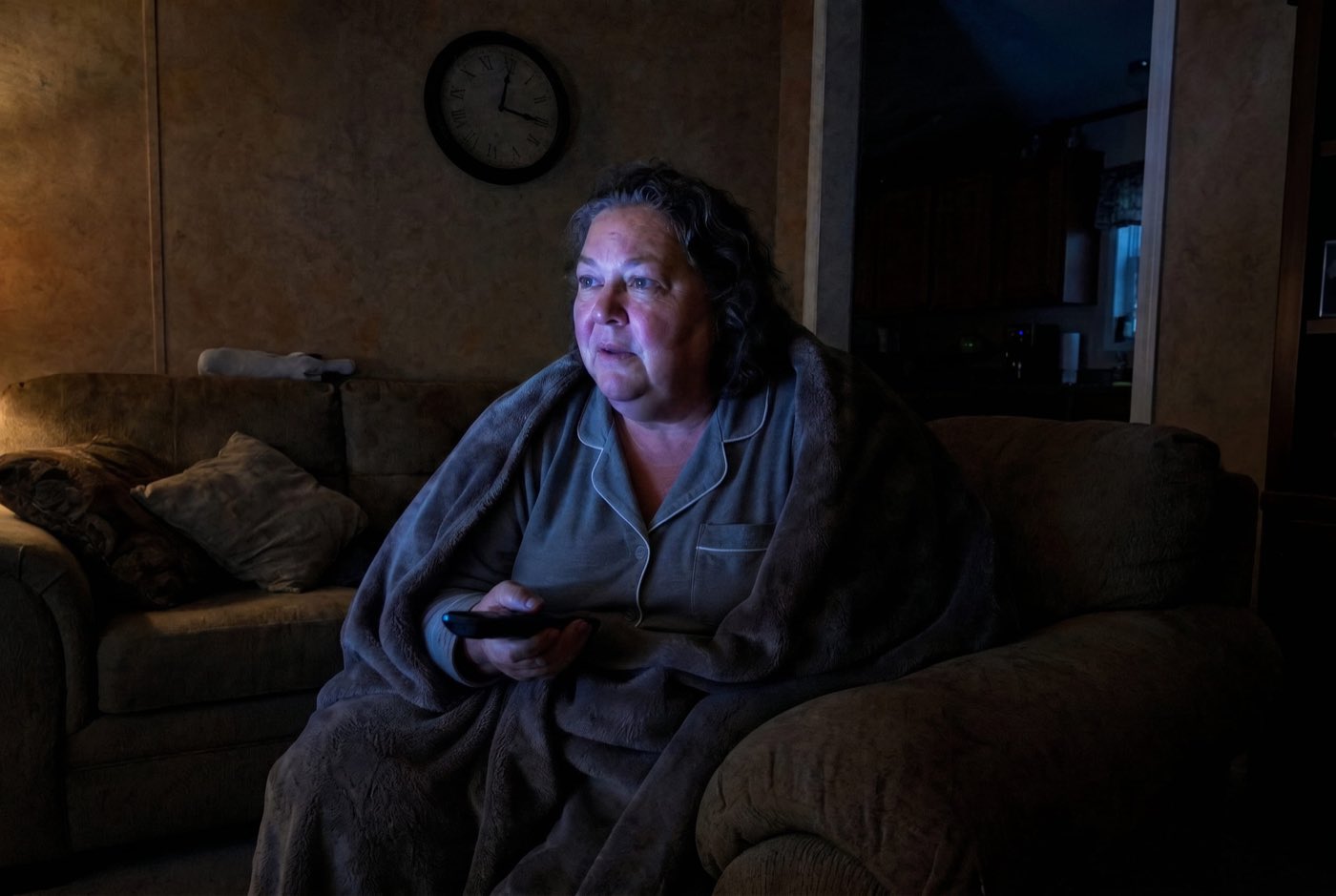 A woman ~55 sitting alone on her living-room couch in the dark at night, wrapped in a soft blanket, lit only by the blue glow of a TV. Her face is tired but suddenly alert and leaning forward, remote in hand, fully caught by what she's watching. A clock faintly reads about 3:00.
